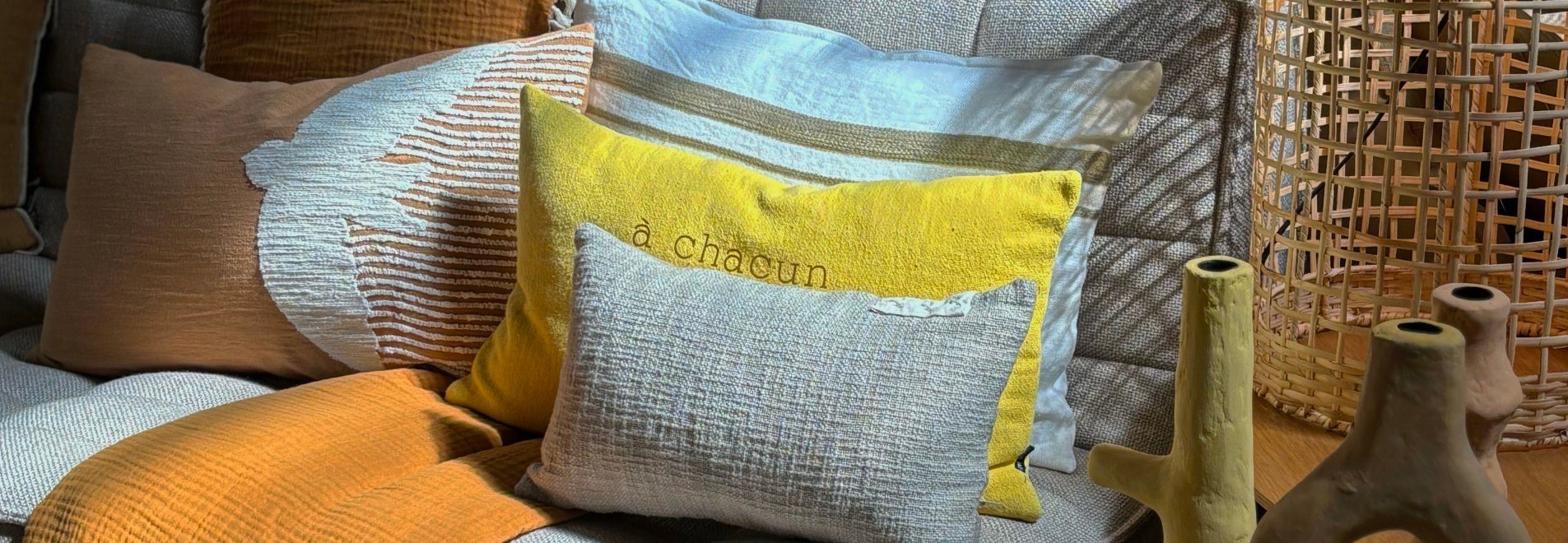 il y  a un jeux de lumière sur un canapé beige ethnicraft, avec plusieurs coussins différentes tailles, matières, motifs et couleurs  en harmonie de style naturel (jaune, beige,oranger)