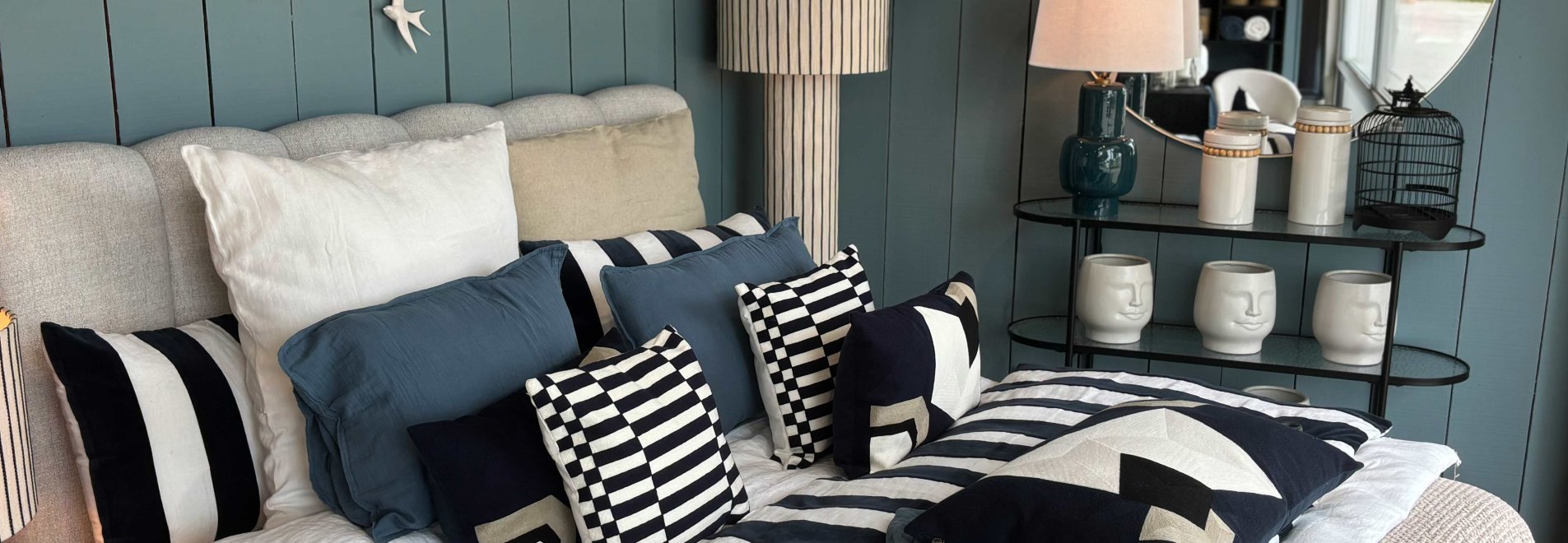 un coin chambre dans les teinte bleu, une tete de lit beige, une lampe à poser avec une grande hauteur à rayure noir et blanche, une variété de coussin et un linge de lit en voile coton